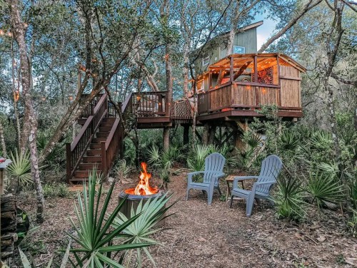 A cozy treehouse surrounded by trees, with a fire pit and two chairs in a serene outdoor setting.