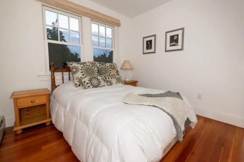 Cozy bedroom with a double bed, decorative pillows, nightstand, lamp, and framed pictures on white walls.