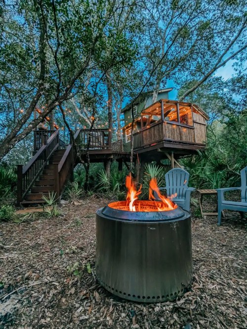 A cozy fire pit in the foreground with a treehouse in the background, surrounded by lush greenery and trees.