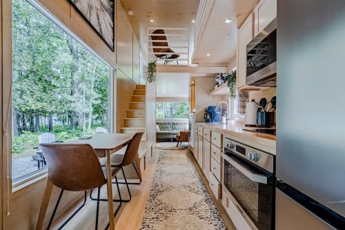 Cozy, modern tiny home interior with a kitchen, dining area, and lofted space, surrounded by greenery.