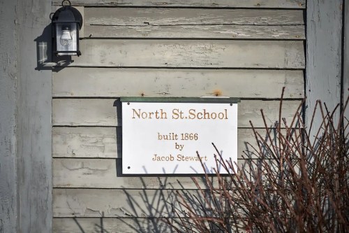 Sign on a wooden wall reading "North St. School, built 1866 by Jacob Stewart," with a lantern above it.