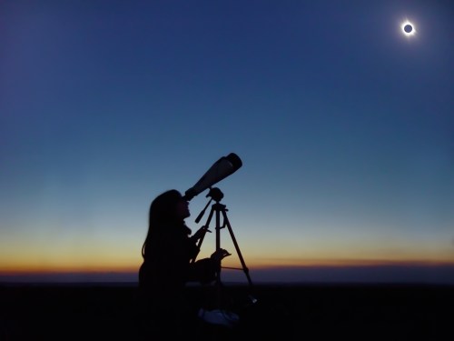 A silhouette of a person using a telescope to observe a solar eclipse against a twilight sky.