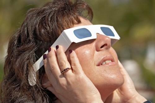 A woman wearing eclipse glasses looks up, shielding her ears with her hands, enjoying a solar event.