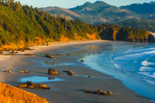 A serene beach scene with gentle waves, rocky shorelines, and lush green hills in the background under a clear sky.