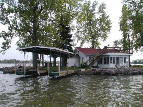 A lakeside house with a red roof, dock, and trees, surrounded by water and rocky shoreline.