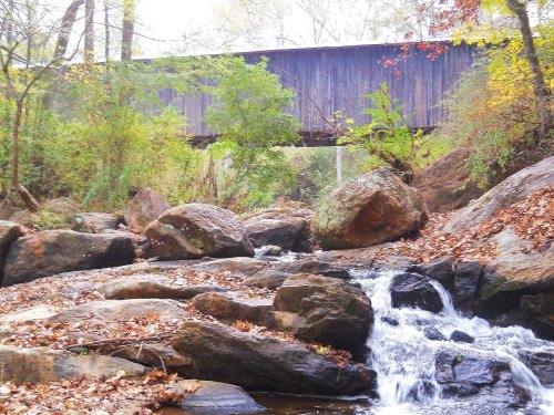 A serene stream flows over rocks, with a wooden bridge above, surrounded by lush greenery and autumn leaves.