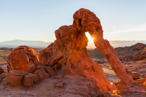 A striking rock formation at sunrise, resembling a natural arch with warm orange hues illuminating the landscape.