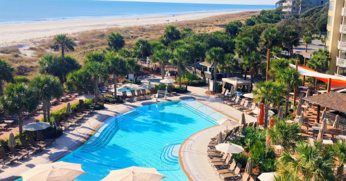 Aerial view of a resort pool surrounded by palm trees, lounge chairs, and a beach in the background.