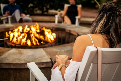 A woman sits by a fire pit, holding a drink, with friends gathered around enjoying the warmth of the flames.