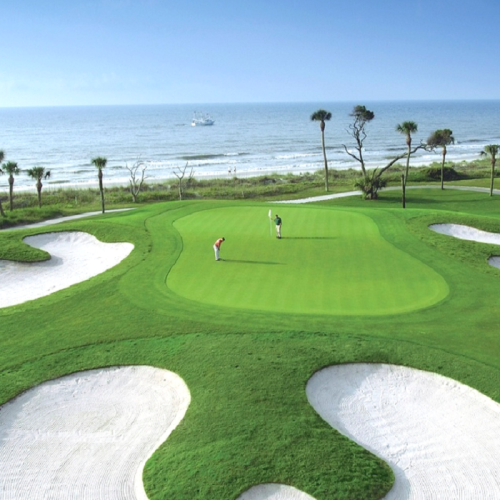 A scenic golf course by the ocean, featuring two golfers on the green and sandy bunkers in the foreground.