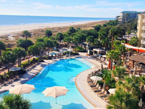 Aerial view of a resort pool surrounded by palm trees, lounge chairs, and a beach in the background under a clear blue sky.