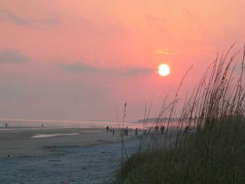 A serene beach at sunset, with silhouettes of people walking and tall grass in the foreground against a colorful sky.