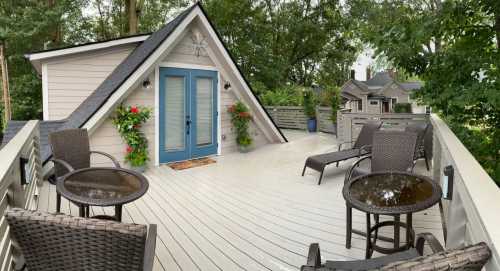 A cozy rooftop deck with two chairs, small tables, and potted plants, surrounded by trees and a charming house.