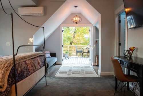 Cozy bedroom with a bed, desk, and a door leading to a deck with outdoor seating and autumn foliage outside.