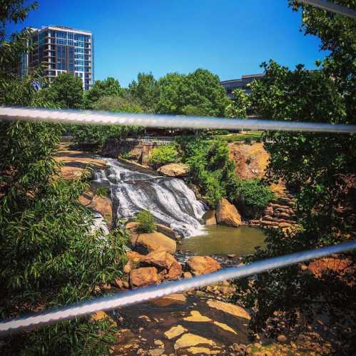 A scenic view of a waterfall surrounded by greenery and buildings under a clear blue sky.