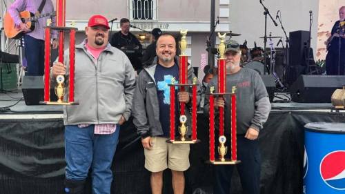 Three men hold large trophies on stage at an outdoor event, with a band performing in the background.