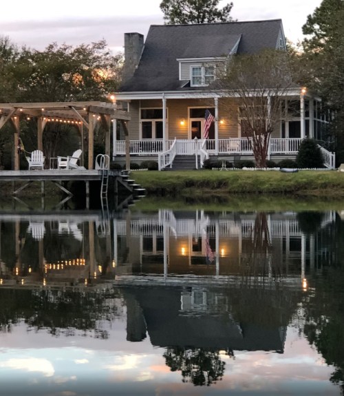 A charming house with a porch and American flag, reflected in a calm lake at sunset, surrounded by trees.