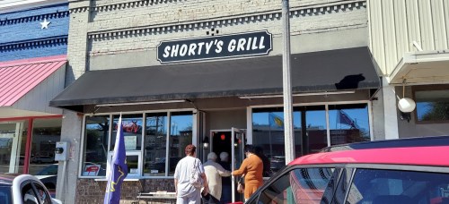 Exterior of Shorty's Grill with a black awning, people entering, and colorful storefronts nearby.