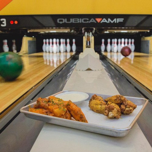 A bowling alley lane with a plate of chicken wings and fries in the foreground, and bowling balls rolling towards pins.