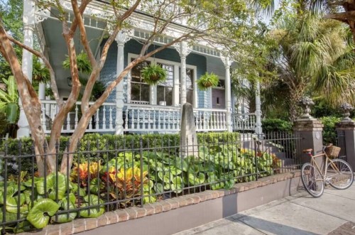 A charming blue house with a porch, surrounded by greenery and flowers, with a bicycle parked nearby.