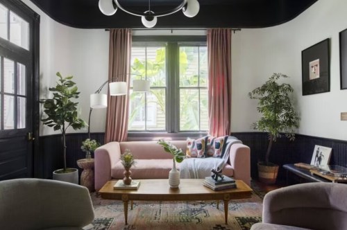 A cozy living room with a pink sofa, plants, a coffee table, and large windows letting in natural light.