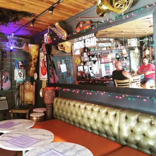 A cozy bar interior with colorful decor, guitars on the walls, and patrons enjoying drinks at a small table.
