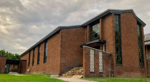 A brick building with large windows, featuring a modern design and a grassy area in front under a cloudy sky.