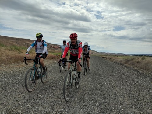 Four cyclists ride on a gravel path surrounded by dry grass and hills under a cloudy sky.