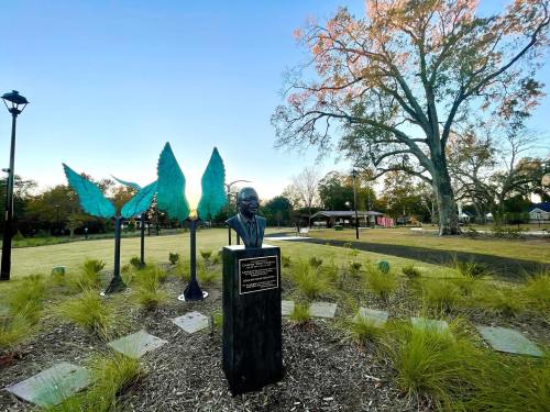 A bust statue surrounded by green leaf sculptures in a park setting with trees and a clear sky.