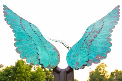 Close-up of a large, turquoise metal sculpture of angel wings against a bright sky.