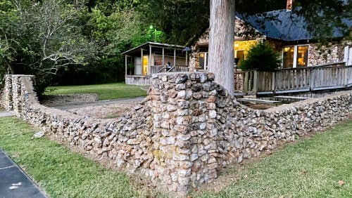 A stone wall surrounds a grassy area in front of a rustic house with large windows and a porch. Trees are nearby.