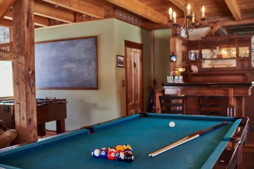 A cozy game room featuring a pool table, foosball table, and a wooden bar with a chalkboard in the background.