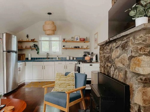 Cozy kitchen with white cabinets, stone fireplace, and a blue tile backsplash, featuring a wooden chair and decorative plants.