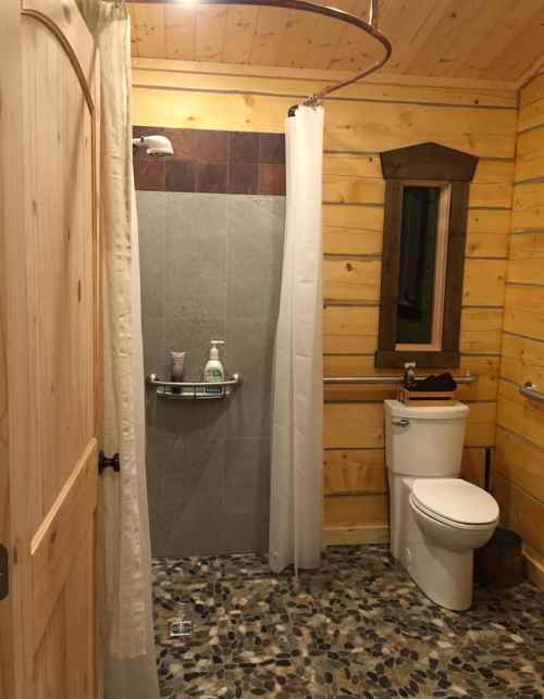 Cozy bathroom with wooden walls, a shower area, toilet, and stone floor. Natural light from a window.