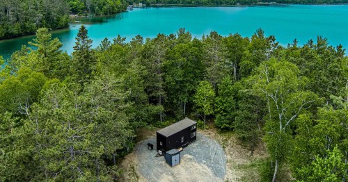 Aerial view of a black cabin surrounded by lush trees near a turquoise lake.