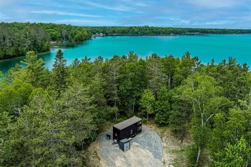 Aerial view of a modern black cabin surrounded by lush trees near a vibrant blue lake.