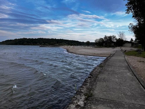 A serene lakeside view with gentle waves, a sandy beach, and a path lined with trees under a cloudy sky.