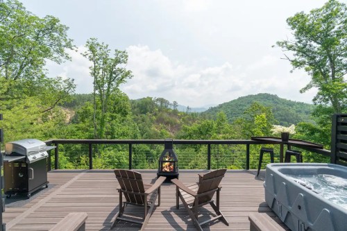 A scenic deck with two chairs, a grill, and a hot tub, overlooking lush green mountains and a cloudy sky.