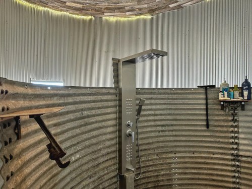 A modern shower with a sleek showerhead inside a circular, corrugated metal enclosure. Shelves hold toiletries.