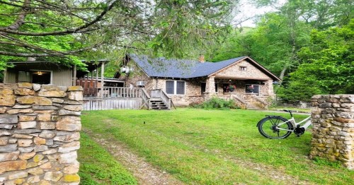 A stone cottage surrounded by greenery, with a bike parked on the grass and a pathway leading to the entrance.