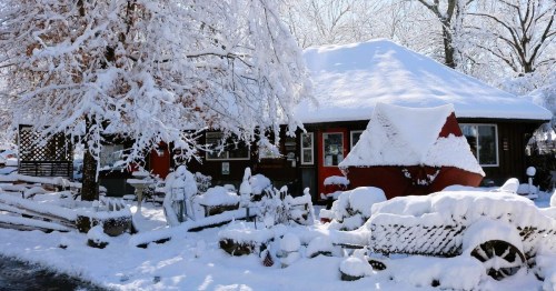 A cozy house covered in snow, surrounded by snow-laden trees and a white landscape, creating a winter wonderland scene.