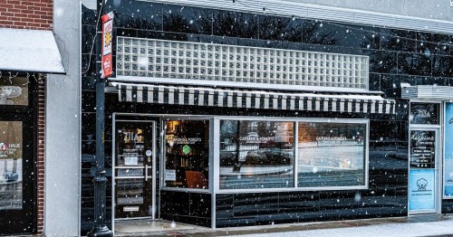 A cozy storefront with large windows, striped awning, and snow falling gently outside.