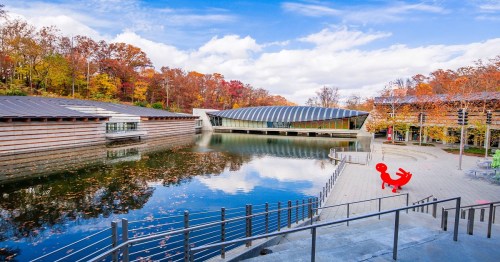 A serene lake reflects colorful autumn trees and modern architecture, with a red sculpture in the foreground.