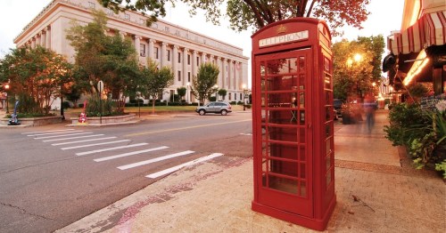 A red telephone booth on a street corner, with a historic building and trees in the background during sunset.