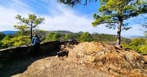 A person stands on a rocky overlook, surrounded by trees, gazing at distant mountains under a blue sky.