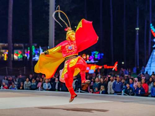 A performer in vibrant red and yellow attire leaps gracefully in front of an audience at a nighttime event.