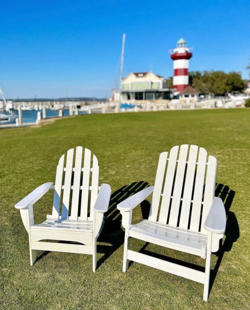Two white Adirondack chairs sit on green grass, with a lighthouse and marina in the background under a clear blue sky.