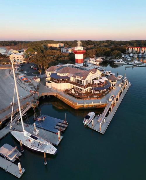Aerial view of a marina with a lighthouse, boats docked, and a waterfront restaurant at sunset.