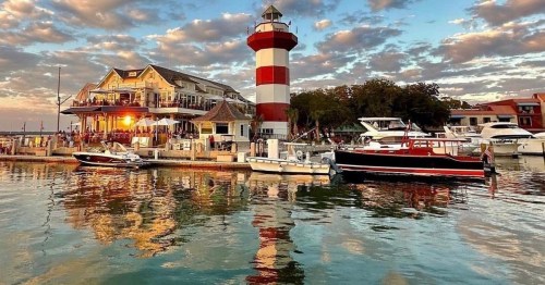A red and white lighthouse beside a waterfront restaurant, with boats docked and colorful clouds reflecting in the water.