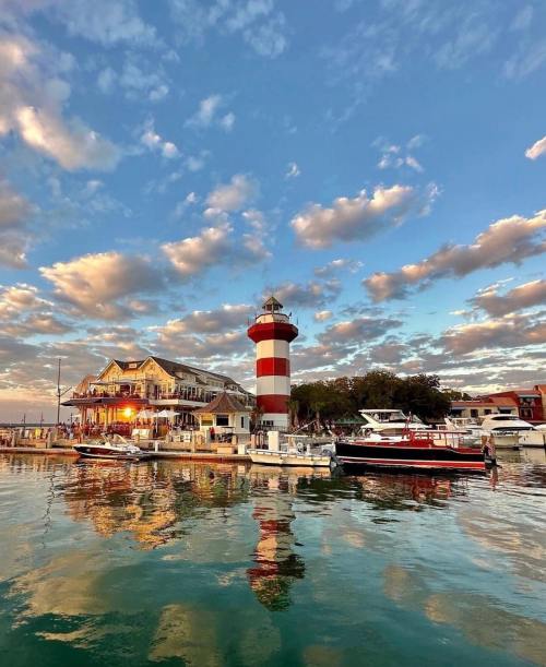 A lighthouse and boats reflect in calm waters under a colorful sky with clouds at sunset.